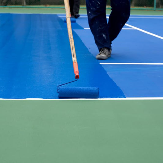 Person painting a tennis court blue with a roller
